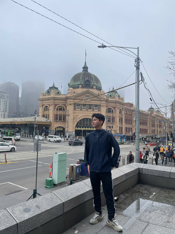 A man wearing a dark navy blue long-sleeve shirt, black pants, and white sneakers stands on a stone ledge in front of a large historic building with a green dome, likely a city landmark, under an overcast sky.